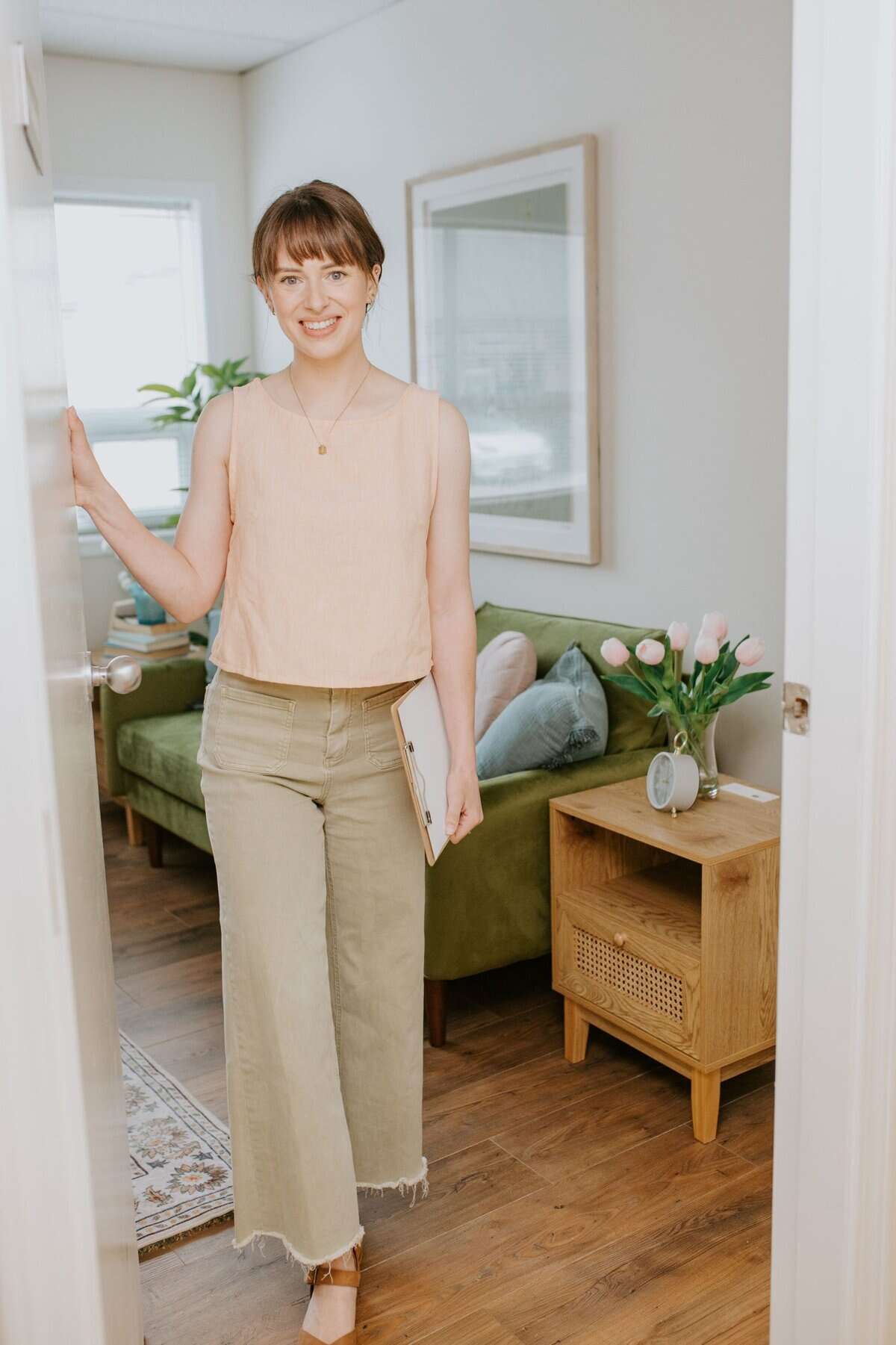 Cozy living room: smiling person, green sofa, tulips, reflecting mirror, minimalist decor.